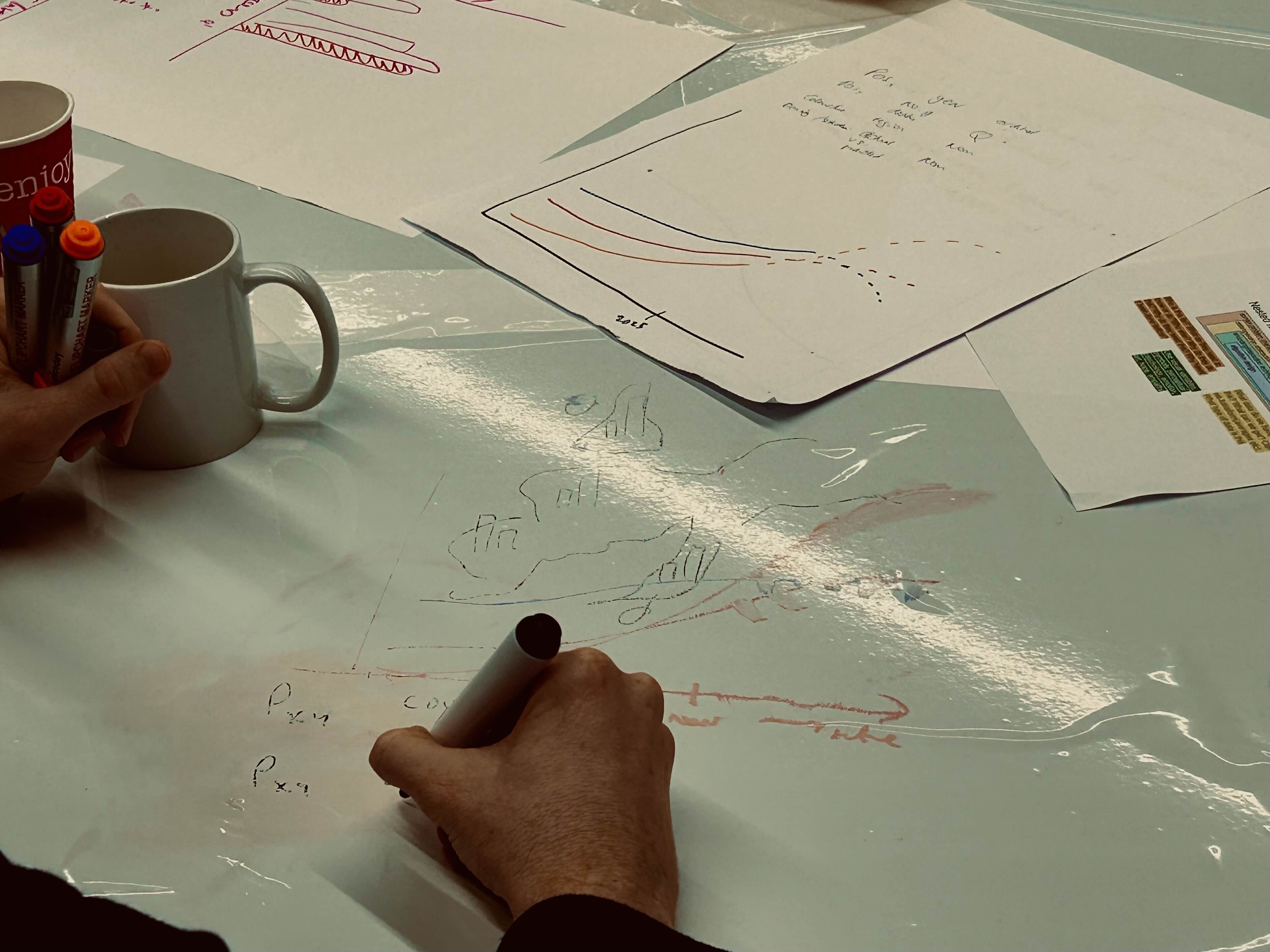 Table with pens, paper and a mug. Human hand is drawing somethig with a marker.